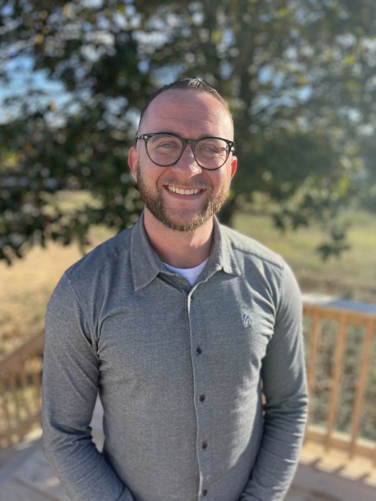 Picture of the salesman James in a nice grey button up shirt, standing outside on a porch a nice sunny day.